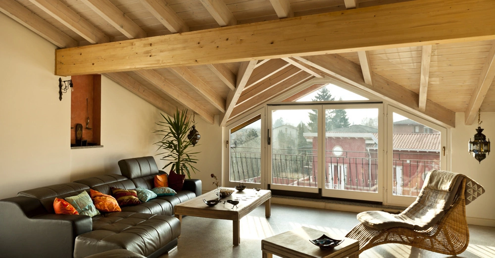 Ledersofa mit bunten Kissen steht vor großen Fenstern in einem Wohnzimmer mit Holzbalkendecke Couchtisch aus Holz daneben Sessel aus Rattan in gemütlicher Ecke mit Pflanzen und Dekorationsgegenständen