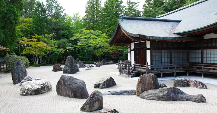 Zen-Garten mit großer Schotterfläche, darauf kleinere Felsbrocken, im rechten Bildrand ein typisch japanischer Holzbau mit geschwungenem Dach und Holzveranda