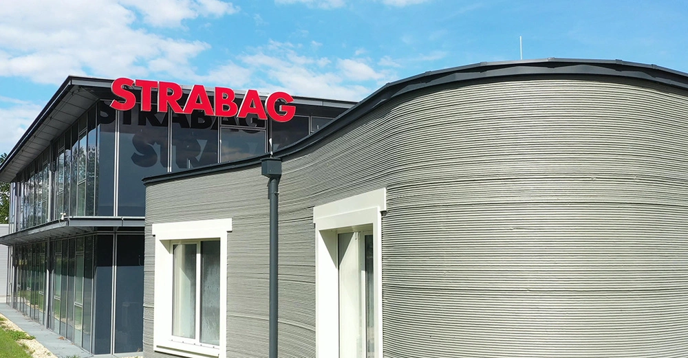 Gebäude mit dem roten Schriftzug STRABAG auf dem Dach. Moderne Architektur mit Glasfassade im Hintergrund graue kurvige Betonwand mit Fenstern im Vordergrund unter blauem Himmel.