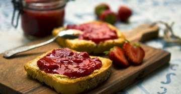 Zwei mit Erdbeermarmelade bestrichene Toastscheiben liegen auf einem Holzbrett neben aufgeschnittenen Erdbeeren, ein verziertes Löffelchen und ein Marmeladenglas stehen im Hintergrund.