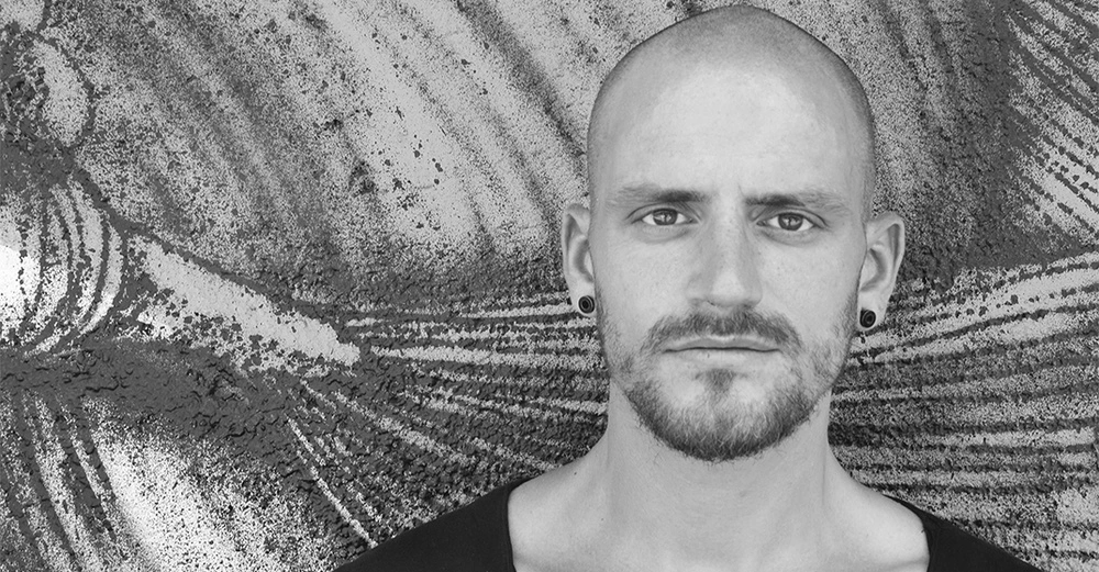 Mann mit Glatze und Bart schaut direkt in die Kamera vor einer strukturierten Wand im Freien. Er trägt schwarze Ohrringe und ein schwarzes Hemd.