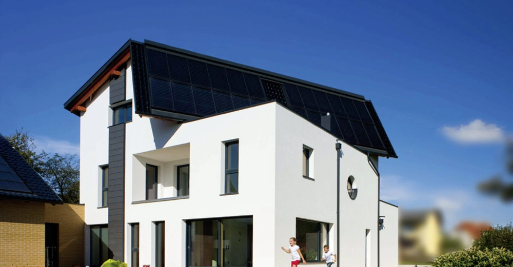Zwei Kinder rennen vor einem modernen weißen Haus mit großen Fenstern und Solarpaneelen auf dem Dach. Das Haus steht in einem gepflegten Garten unter blauem Himmel.
