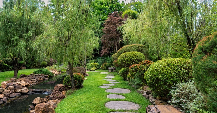 Steinweg führt durch gepflegten Garten mit Teich links und Sitzbank rechts umgeben von üppigen Sträuchern und Bäumen im Hintergrund grün und bewölkt.