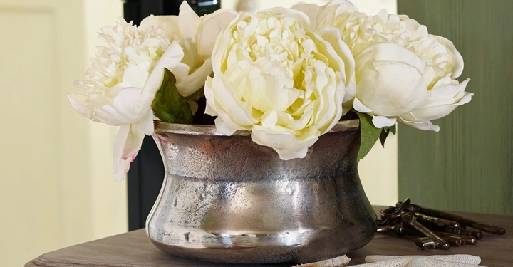 Galerie-Bild 8: Metallvase mit weißen Pfingstrosen steht auf einem Holztisch neben Seesternen und alten Schlüsseln in einem wohnlichen Innenraum.