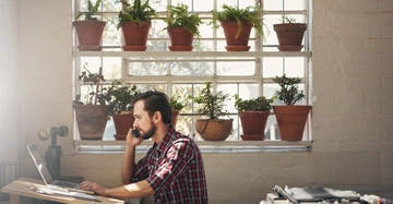 Mann telefoniert sitzend am Schreibtisch vor Laptop helle Werkstatt mit großen Fenstern und vielen Topfpflanzen auf Fensterbänken Sonnenlicht durchflutet den Raum Skizzierungen und Werkzeuge auf dem Tisch