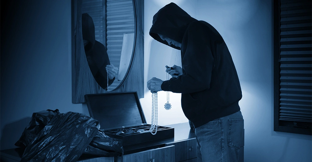 Eine Person im Kapuzenpullover untersucht Schmuck aus einem offenen Safe in einem Raum mit einem Spiegel an der Wand und geschlossenen Jalousien am Fenster.