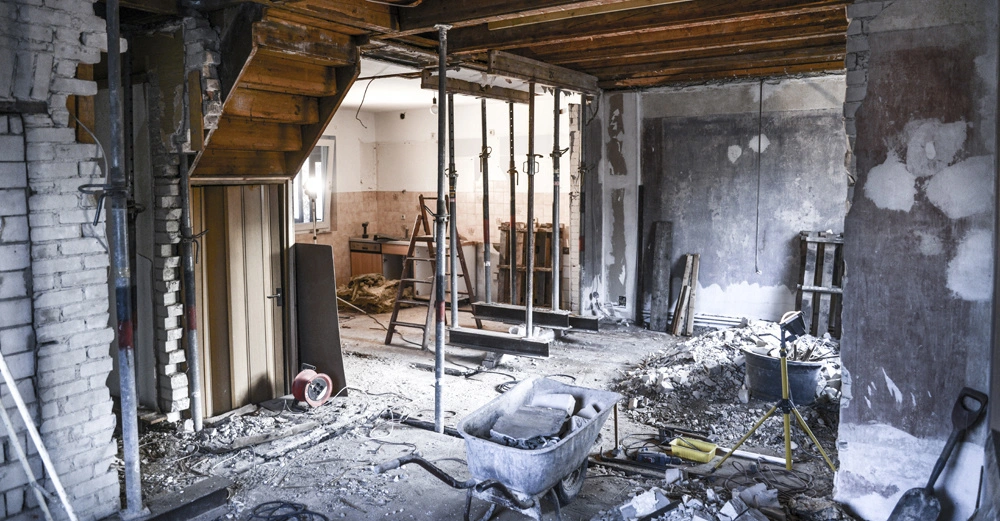 Baustelle im Haus zeigt einen Abbruch mit Schubkarre in der Mitte, Ziegelsteine und Staub auf dem Boden, Stützpfosten, Leiter und Werkzeuge in einem teilweise entkernten Raum.
