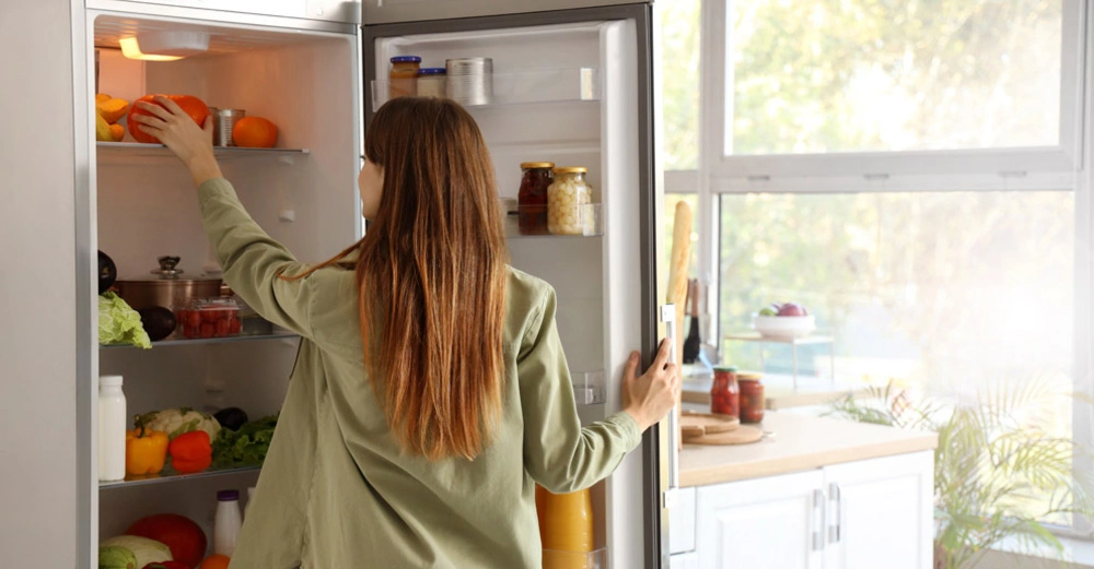 Frau in grüner Bluse und mit langen rotbraunen Haaren nimmt etwas aus dem geöffneten Kühlschrank. Draußen scheint die Sonne.