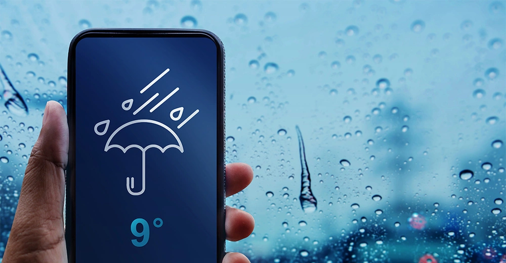 Eine Hand hält ein Smartphone das eine Wetter-App mit einem Regenschirm-Symbol und der Temperatur 9 Grad anzeigt im Hintergrund regnet es.