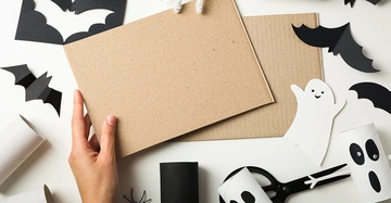 Eine Hand hält ein Stück Pappe in einem Bastelumfeld mit ausgeschnittenen schwarzen Fledermäusen und weißen Gespenstern auf einem weißen Tisch.