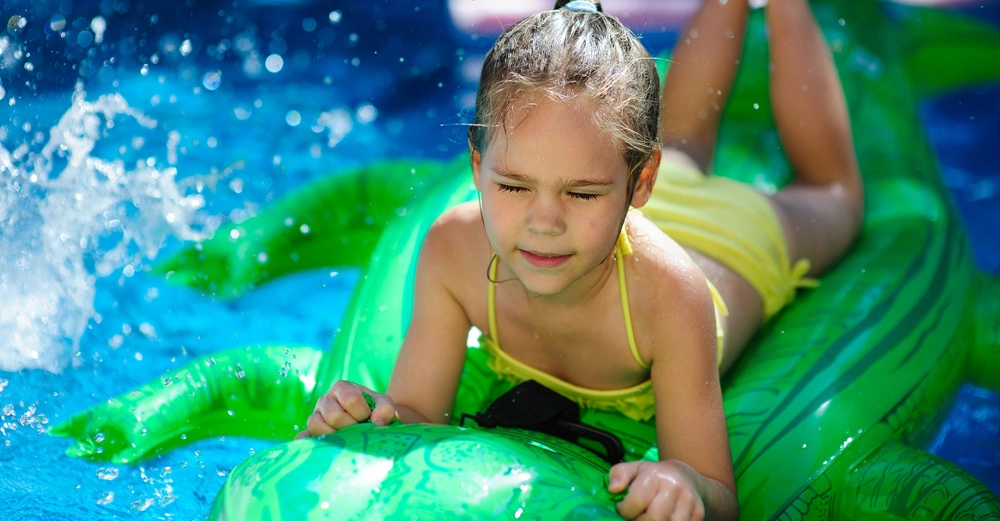 Mädchen liegt auf aufblasbarem Krokodil im blauen Pool spritzt Wasser herum.
