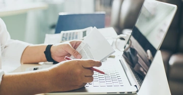 Hände halten ein Dokument und einen roten Stift Arbeiten an einem Laptop auf einem Schreibtisch im Büro mit einem Taschenrechner und Papieren im Hintergrund.