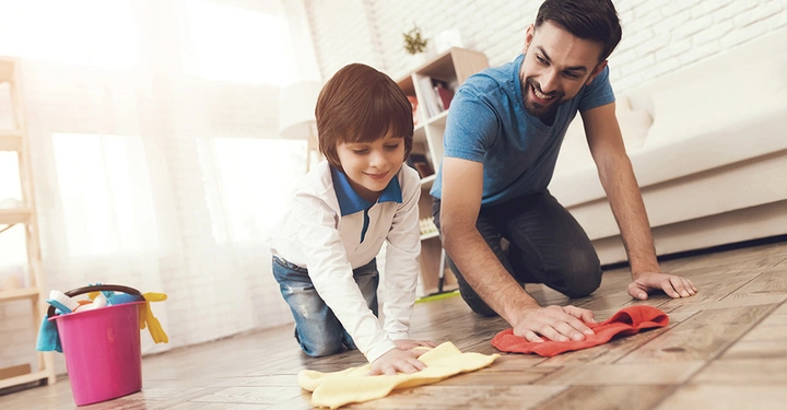 Ein Mann und ein Kind reinigen gemeinsam den Fußboden mit Tüchern in einem hellen Wohnzimmer. Im Hintergrund steht ein pinker Eimer mit Reinigungsutensilien.