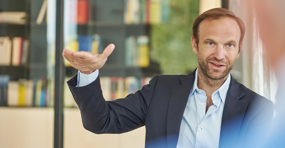 Mann trägt Anzug gestikuliert mit erhobener Hand steht in einem Raum mit Bücherregalen im Hintergrund.