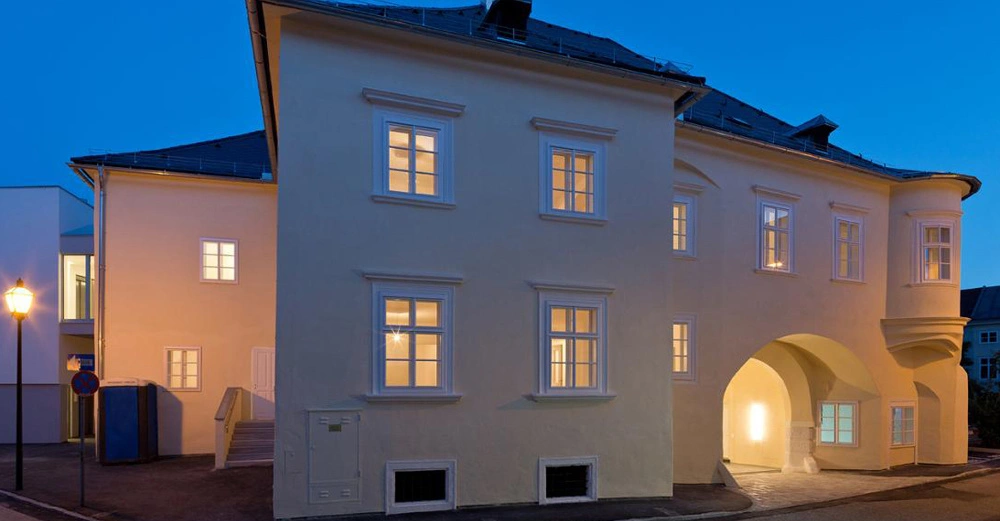Ein großes weißes Gebäude mit beleuchteten Fenstern steht in einer städtischen Umgebung bei Dämmerung. Eine Straßenlaterne beleuchtet den Gehweg daneben.