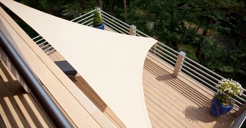 Blick von oben auf Holzterrasse mit Metallgeländer und beiges, großes Sonnensegel, das für Schatten sorgt. Wald im Hintergrund.