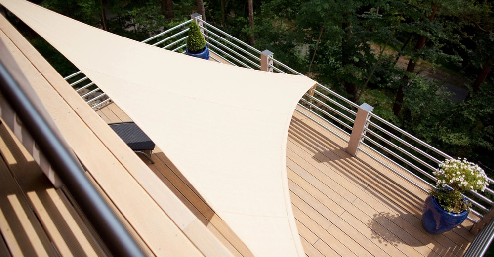 Blick von oben auf Holzterrasse mit Metallgeländer und beiges, großes Sonnensegel, das für Schatten sorgt. Wald im Hintergrund.