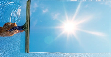 Hand hält Fensterabzieher wischt ein Fenster sauber ein sonniger Himmel mit ein paar Wolken im Hintergrund Reinigung zeigt klare Unterschiede zwischen sauberen und schmutzigen Bereichen