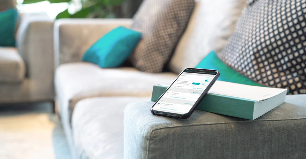 Smartphone liegend auf einem Buch auf einem grauen Sofa in einem Wohnzimmer mit bunten Kissen im Hintergrund Text auf dem Smartphone Display ist teilweise lesbar zeigt eine App Oberfläche