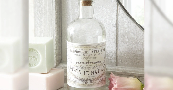 Galerie-Bild 7: Eine gläserne Flasche mit Korkverschluss und dem Etikett Parfumerie Extra-Fine Maison Fondée en 1818 Paris-Bruxelles Savon Le Naturel steht neben zwei Seifenstücken vor einem gerahmten Spiegel auf einer weißen Oberfläche mit einer rosa Blume.