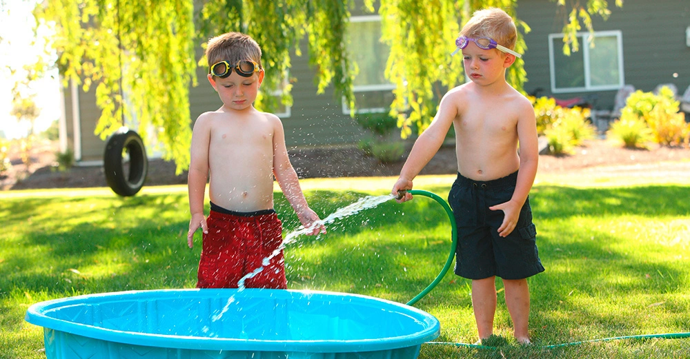 2 blonde Jungs in der Badehose stehen im Garten und befüllen ein kleines Schwimmbecken mit Wasser aus dem Gartenschlauch