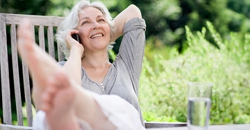 Ältere Frau mit grauen Haaren aber sehr jugendlich sitzt mit Beinen auf dem Tisch telefonierend im Garten. 
