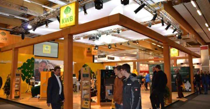Galerie-Bild 21: Menschen gehen an einem Messestand mit der Aufschrift KWB vorbei Messehalle Holzstruktur, Girlandenlichter und technische Produkte im Hintergrund.