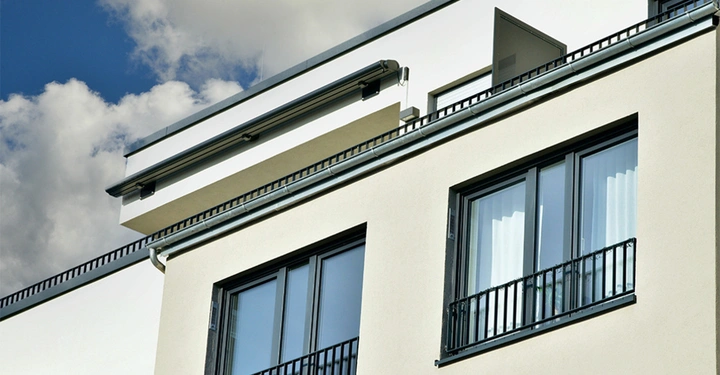 Ein modernes mehrstöckiges Gebäude zeigt Fenster mit Geländern im Vordergrund. Das Gebäude hat eine glatte weiße Fassade und steht unter einem bewölkten Himmel.