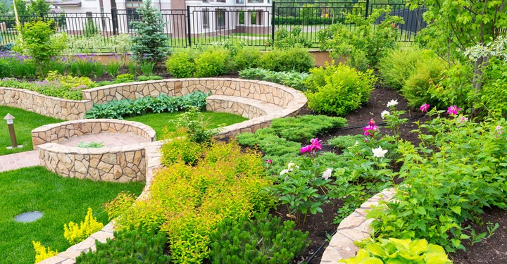 Steingartenmauer mit integrierten Sitzplätzen umgibt grüne Pflanzen und bunte Blumen in einem gepflegten Garten vor einem Wohngebäude mit Zaun im Hintergrund