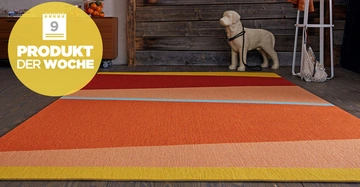 Bunter Teppich liegt auf Holzboden vor einer Holzwand mit dekorativer Hundeskulptur und einem Seitentisch Text: 9 PRODUKT DER WOCHE