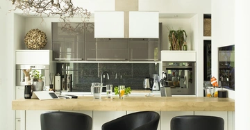 Moderne Küche mit Holzarbeitsplatte drei schwarzen Barhockern und grauer Rückwand eine Kaffeekanne und Gläser auf der Theke Pflanzen und Küchengeräte umgeben von weißen Schränken.