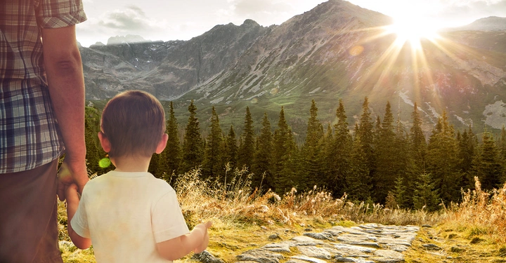 Ein Kind hält die Hand eines Erwachsenen und blickt auf einen sonnigen Bergpfad mit Nadelbäumen und Gipfeln im Hintergrund.