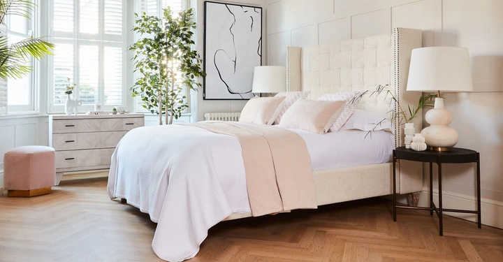 Beige Bett mit rosa Decke und Kissen steht in einem hellen Schlafzimmer mit Holzboden eine Kommode steht neben großen Fenstern Zimmerpflanzen und Kunstwerk an den Wänden und eine Tischlampe auf dem Nachttisch