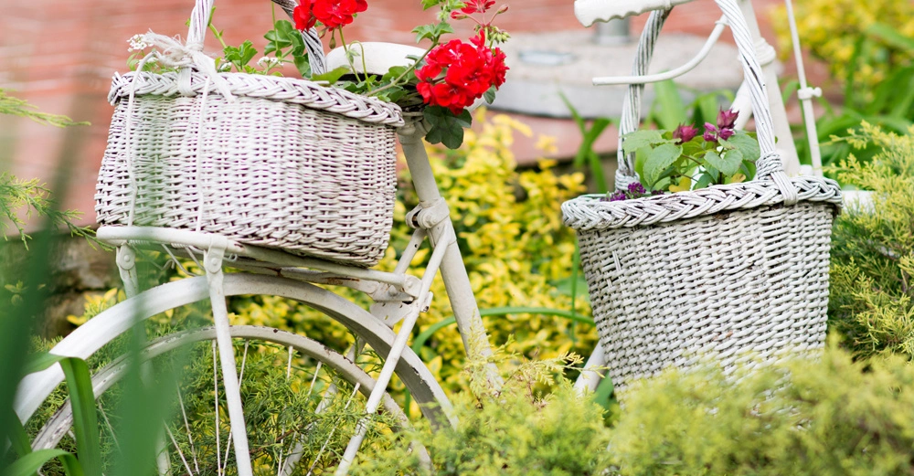 Altes weißes Fahrrad mit zwei Körben voller Blumen steht dekorativ im Garten zwischen grünen Sträuchern und bunten Blumen vor einem roten Backstein-Hintergrund.