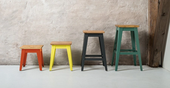 Vier Hocker stehen still in aufsteigender Größe aufgefächert in einer Reihe auf einem Boden neben einer braunen Wand und einem hölzernen Stützbalken.