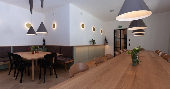 Galerie-Bild 14: Holztische stehen in einem modernen Essbereich mit Hängelampen und dekorativen Pflanzenvase. Im Hintergrund befinden sich eine Sitzbank und eine Rückwand mit rundem Lichtdesign.