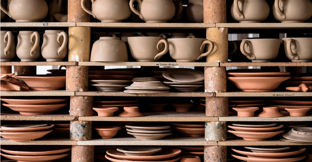 Stilleben: Regal voll gestellt mit zahlreichen Töpfen, Schüsseln, Tellern und Kannen aus Ton und Keramik. Noch unglasiert und ungebrannt. Alles erscheint rötlich, beige und braun. 