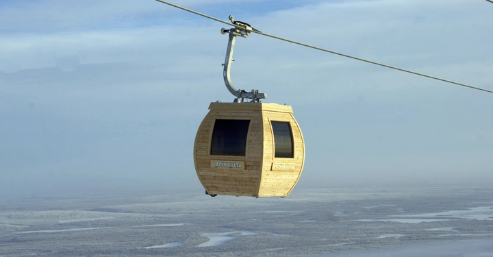Eine hölzerne Seilbahn-Gondel fährt in der Luft über eine schneebedeckte Landschaft. Auf der Gondel steht der Text "LAPIN KULTA".