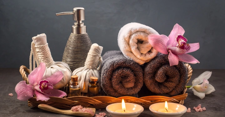 Ein Korb mit Handtüchern und Blüten steht auf einer dunklen Oberfläche umgeben von Kerzen und Spa-Zubehör in einer ruhigen Umgebung.