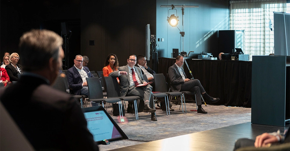Personen sitzen auf Stühlen in einem Konferenzraum und hören einem Vortrag zu. Ein Redner spricht auf der Bühne. Der Raum ist modern eingerichtet und von Technikgeräten umgeben.