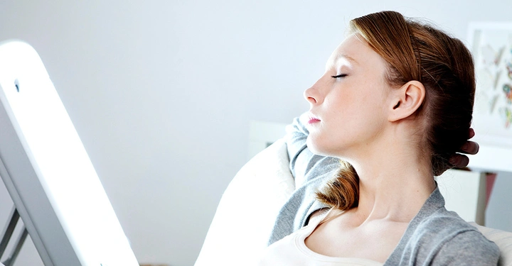 Eine entspannte Frau sitzt vor einer hellen Tageslichtlampe zur Lichttherapie-Behandlung gegen Winterdepression und saisonale Verstimmung.