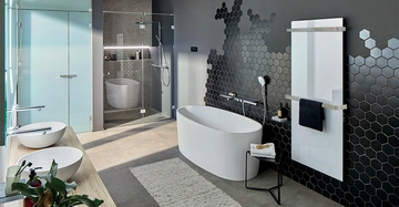 Freistehende Badewanne steht in einem modernen Badezimmer mit schwarzer Wabenfliesenwand und begehbarer Glasdusche im Hintergrund zwei Waschbecken auf einer Holzablage und eine Zimmerpflanze in der Ecke.