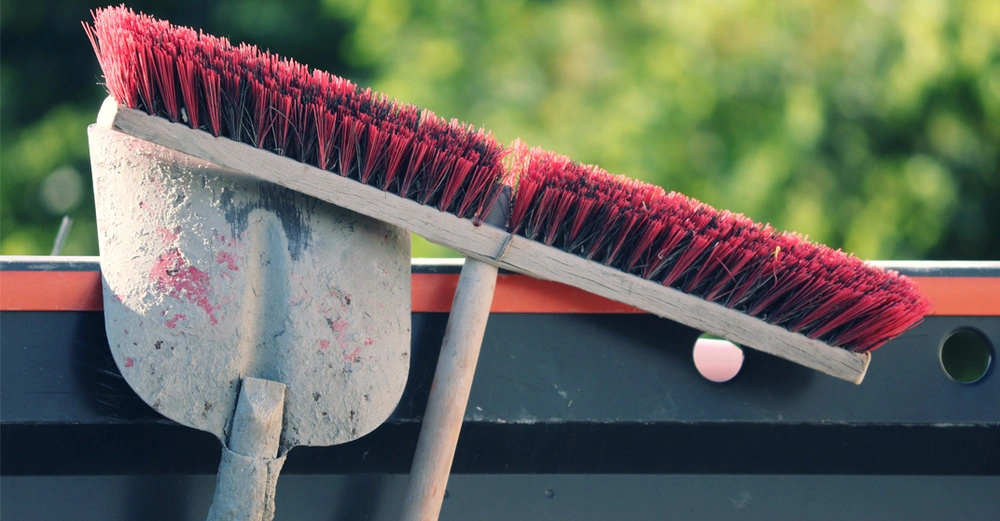Eine dreckige Schaufel und ein Besen mit roten Borsten lehnen an einem schwarzen Container.