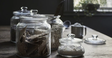 Glasbehälter mit Deckeln stehen auf einem Holztisch und enthalten Lebensmittel wie Kekse Haferflocken und Mehl Im Hintergrund sind ein Fenster mit Pflanzen und eine Schaufel zu sehen