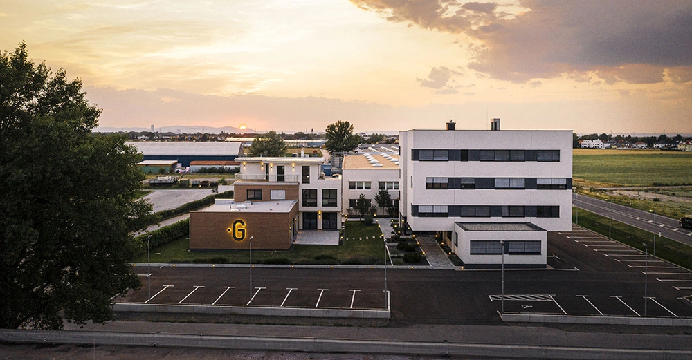 Zwei moderne Gebäude mit großen Fenstern stehen nebeneinander in einem weitläufigen, leeren Parkplatz, umgeben von Feldern im Hintergrund bei Sonnenuntergang. Großes gelbes G auf einem der Gebäude.