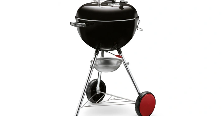 Galerie-Bild 2: Ein schwarzer Kugelgrill steht auf einem Stativ mit zwei Rädern und einem Griff. Ein Ascheauffangbehälter hängt darunter. Text Weber Original Kettle.