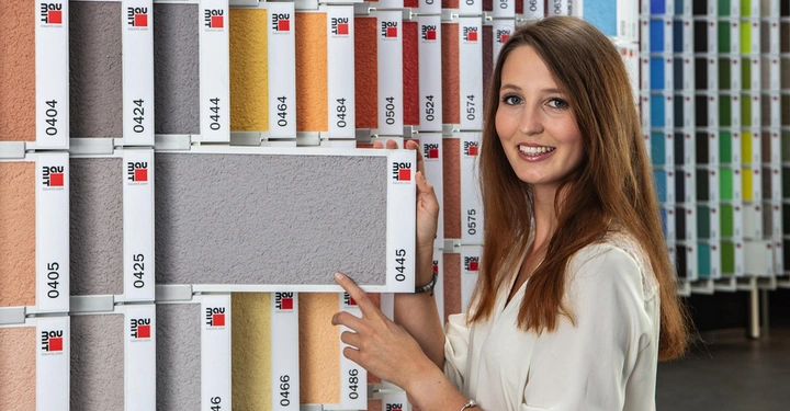 Galerie-Bild 2: Frau hält eine graue Farbprobe mit der Nummer 0445 in einem Farbstudio umgeben von verschiedenen Farbmustern mit Nummern wie 0404 0424 0444 0464.