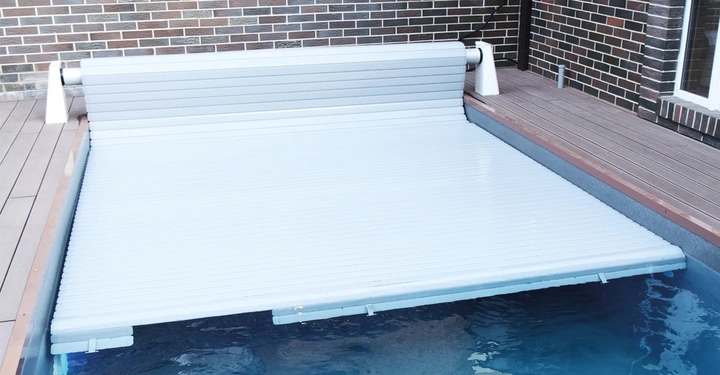 weiße stabile Rollabdeckung für Pool, halb ausgefahren, darunter Poolwasser zu sehen, rundum braune Poolsteine