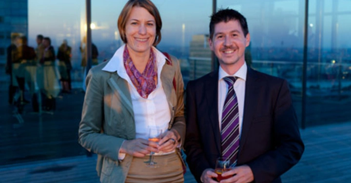 Galerie-Bild 20: Zwei Personen halten Gläser mit Getränken und lächeln in die Kamera. Sie stehen auf einer Terrasse eines modernen Gebäudes in einer Stadtlandschaft bei Sonnenuntergang vorhanden.