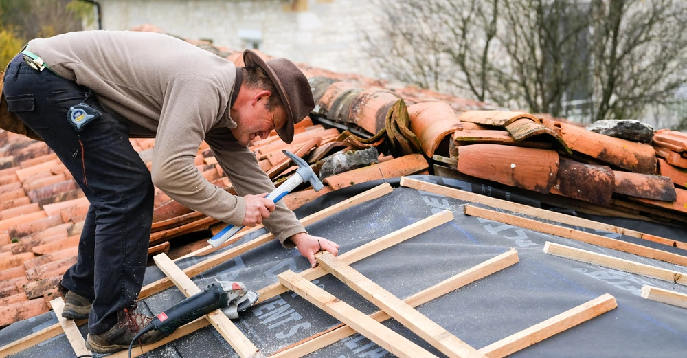 Arbeiter nagelt Dachlatten an teilweise abgedecktem Hausdach fest. Auf dem Dach hinten liegen abgedeckte, gestapelte Dachziegel in orange.